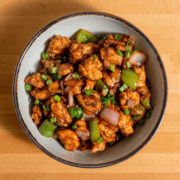 A bowl of General Tso's chicken, with green peppers, onions, and scallions, on a wooden table.